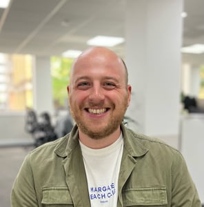 Ollie is a white man in his twenties with a beard, wearing a white t shirt and green shirt, smiling at the camera.