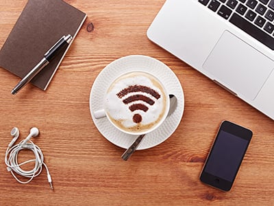 a wifi symbol is painted in chocolate on the milk foam of a coffee cup, which sits squarely between a laptop, phone, headphones and a notebook