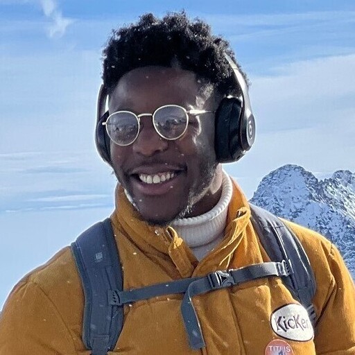 A headshot of Dean Akinwale, a man wearing glasses and headphones, in a cold climate with snowy mountains in the background