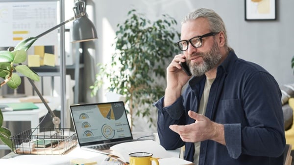 Man with gray hair in ponytails and beard talks on phone with report showing on laptop