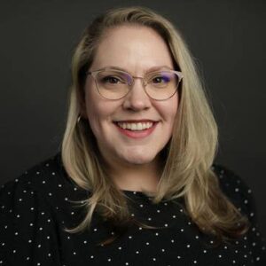 A headshot of Bethany Nothstein, a blonde woman wearing glasses and a black blouse with small white polka dots.