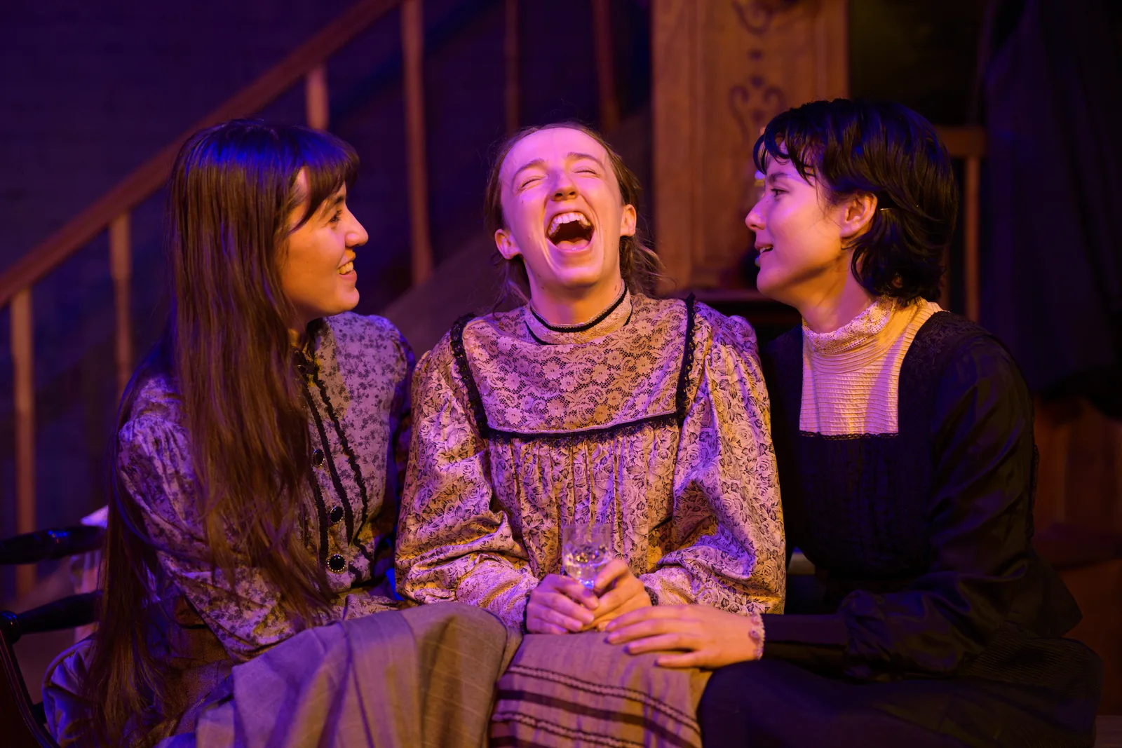Three Women Sit Closely Together As The Central Woman Laughs And The Others Lean Toward Her.