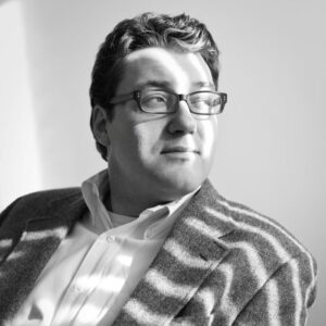 A photo of Jack Wright wearing a button-down, glasses, and a blazer, in greyscale.