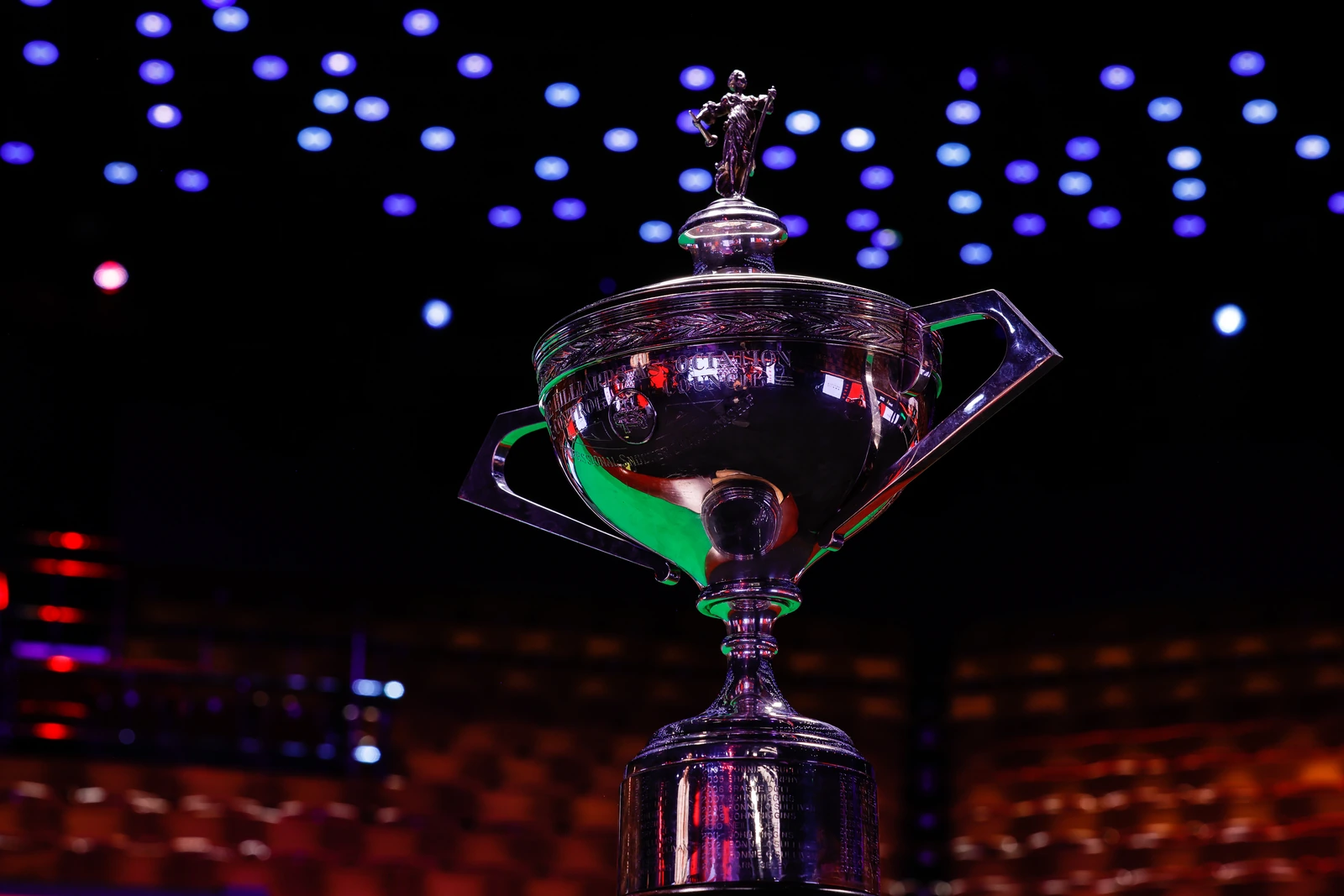 Close-Up Of The Silver World Snooker Championship Trophy Beneath Purple And Blue Arena Lights, With Seating Blurred Behind It.