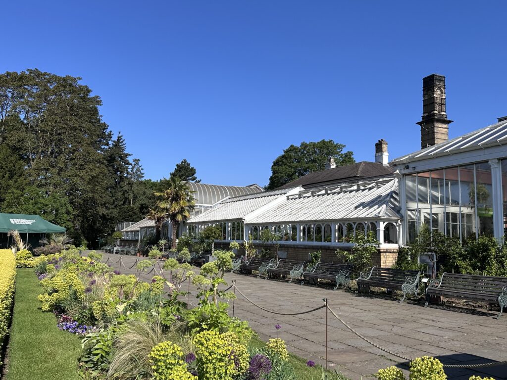 Exterior View Of A Conservatory At Birmingham Botanical Gardens In Summer, With Benches For Visitors And Flowers In Bloom.